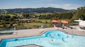 Außenpool im AIGO welcome family mit Badenden auf einer Terrasse, dahinter grüne Hügellandschaft und Felder unter blauem Himmel.