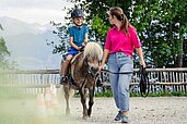 Junge sitzt auf einem Pony und wird von einer Reitlehrerin im Allgäuer Berghof geführt.