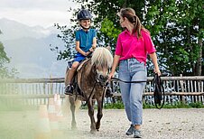 Junge sitzt auf einem Pony und wird von einer Reitlehrerin im Allgäuer Berghof geführt.