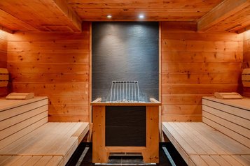 Holzverkleidete Sauna mit zwei Sitzbänken links und rechts sowie einem Saunaofen in der Mitte vor einer dunklen Steinwand im Familienhotel Amiamo.