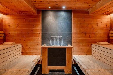 Holzverkleidete Sauna mit zwei Sitzbänken links und rechts sowie einem Saunaofen in der Mitte vor einer dunklen Steinwand im Familienhotel Amiamo.