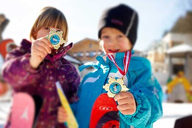 Zwei stolze Kinder im Winteroutfit präsentieren ihre gewonnenen Medaillen beim Skifahren, symbolisch für die kinderfreundlichen Wintersportaktivitäten im Familienhotel Amiamo im Salzburger Land.