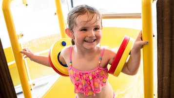 Ein Mädchen mit Schwimmflügeln freut sich am Ende einer gelben Wasserrutsche im Familienhotel