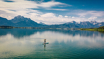 Ein rießiger See mit Bergpanorama im Hintergrund. Auf dem See paddelt eine Frau und genießt die Natur.