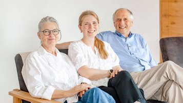 Drei Personen aus der Gastgeberfamilie Hörl – zwei ältere Erwachsene und eine junge Frau – sitzen entspannt nebeneinander auf einem Sofa und lächeln freundlich.