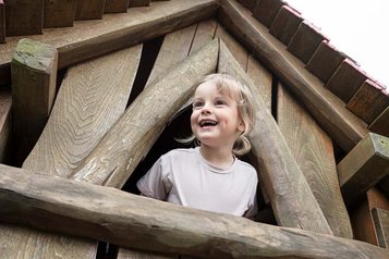 Ein fröhliches Kind schaut aus dem Fenster eines Holzkletterturms am Spielplatz eines Familotel.