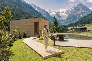 Ein Steg führt um einen kleinen Naturteich zur außenliegenden Sauna, im Hintergrund Berge beim Hotel Bella Vista in Trafoi in Südtirol.