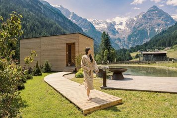 Ein Steg führt um einen kleinen Naturteich zur außenliegenden Sauna, im Hintergrund Berge beim Hotel Bella Vista in Trafoi in Südtirol.
