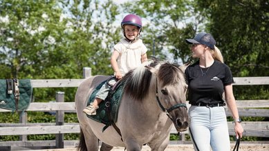 Kind reitet lachend auf einem Pony während des Reitunterrichts in der Familotel Erlebniswelt Reiten.