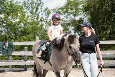 Kind reitet lachend auf einem Pony während des Reitunterrichts in der Familotel Erlebniswelt Reiten.