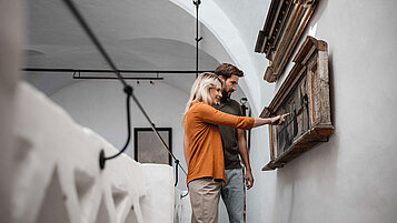 Ein kulturbegeistertes Paar bestaunt Werke an einer Wand in einem Museum in Südtirol. Eine super Gelegenheit im Familienurlaub sich treiben zu lassen und sich nachher mit der Familie auszutauschen.