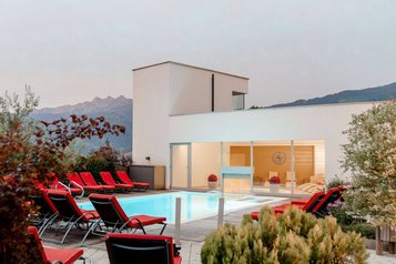 Beleuchteter Außenpool des Familienhotels Amiamo am Abend mit vielen roten Liegen, Glasfront zur Relax-Lounge und Bergen im Hintergrund.