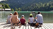 Vater und Mutter sitzen gemeinsam mit ihren drei Kindern auf dem Steg des Königssees und blicken aufs Wasser.