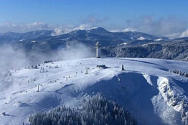 Winterliche Landschaft mit Schneedecke und Skiliften am Feldberg im Schwarzwald, mit einem Turm, der über die Nebelschwaden ragt.