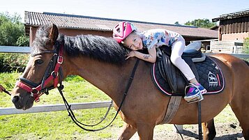 Glückliches Mädchen sitzt auf dem Reitplatz des Landhuus Laurenz auf einem Pferd und kuschelt mit dem Tier.