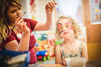 Kinderbetreuerin im Happy-Club des Familienhotels Landgut Furtherwirt bemalt das Gesicht eines Mädchens mit bunten Farben.