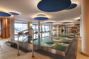 Großzügiger Innenpool im Zauchenseehof mit modernen Deckenelementen in Blautönen, stimmungsvoller Beleuchtung und Liegen entlang der Panorama­fenster.