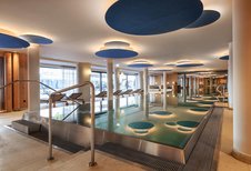 Großzügiger Innenpool im Zauchenseehof mit modernen Deckenelementen in Blautönen, stimmungsvoller Beleuchtung und Liegen entlang der Panorama­fenster.