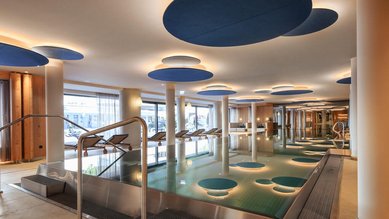 Großzügiger Innenpool im Zauchenseehof mit modernen Deckenelementen in Blautönen, stimmungsvoller Beleuchtung und Liegen entlang der Panoramafenster.