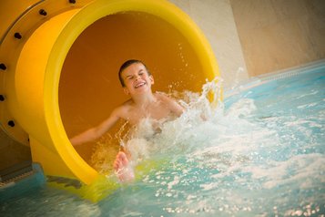 Junge mit strahlendem Lächeln rutscht aus einer gelben Wasserrutsche ins Becken des Landguts Furtherwirt und sorgt für spritzigen Badespaß
