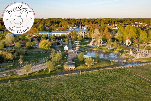 Familien Wellness Hotel Seeklause an der Ostsee mit großem Abenteuerspielplatz, Teich und Holzstegen, umgeben von Grünflächen und Ortschaft im Hintergrund.