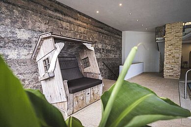 Originelle Sitzgelegenheit in Form eines rustikalen Holzhauses im Innenbereich eines Familienhotels, vor einer Wand mit natürlicher Steinoptik.