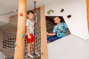 Ein Junge steht auf einem Balancierseil an einem Kletterbaum, ein anderer sitzt in einem Spielversteck mit Klettergriffen.
