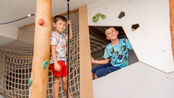 Ein Junge steht auf einem Balancierseil an einem Kletterbaum, ein anderer sitzt in einem Spielversteck mit Klettergriffen.