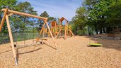Spielplatz mit Klettergerüst, Nestschaukel und Karussell aus Holz im Familienhotel Sonnenhügel in der Rhön, umgeben von Bäumen und unter blauem Himmel.