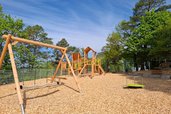 Spielplatz mit Klettergerüst, Nestschaukel und Karussell aus Holz im Familienhotel Sonnenhügel in der Rhön, umgeben von Bäumen und unter blauem Himmel.