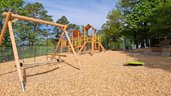 Spielplatz mit Klettergerüst, Nestschaukel und Karussell aus Holz im Familienhotel Sonnenhügel in der Rhön, umgeben von Bäumen und unter blauem Himmel.