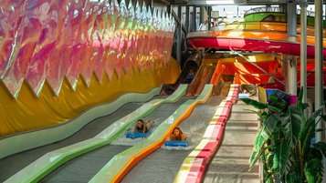 Mehrbahn-Wasserrutsche in einer farbenfrohen Indoor-Anlage, auf der mehrere Kinder auf Matten nebeneinander ins Becken gleiten, umgeben von leuchtenden Paneelen und tropischen Pflanzen.