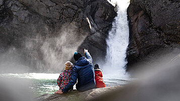 Vater sitzt mit seinen beiden Kindern vor einem Wasserfall in Allgäu und genießt die Aussicht.