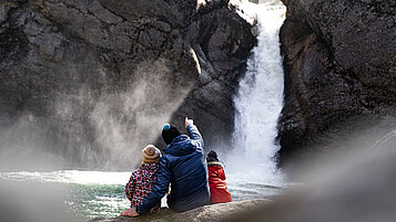 Vater sitzt mit seinen beiden Kindern vor einem Wasserfall in Allgäu und genießt die Aussicht.