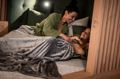 Mutter und Kind kuscheln in Bademänteln unter einer Decke im Wellnessbereich des Familienhotel Alpenhof Dolomit Family.