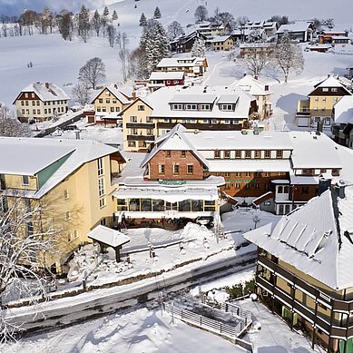 Luftaufnahme einer verschneiten Hotelanlage im Schwarzwald, umgeben von malerischen Häusern und schneebedeckten Bäumen, unterstrichen durch die idyllische Winterstimmung.