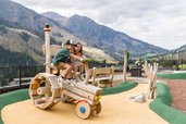 Zwei Kinder spielen auf einem Holztraktor am Abenteuerspielplatz des Familienhotels Almfamilyhotel Scherer mit Bergkulisse im Hintergrund.