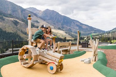 Zwei Kinder spielen auf einem Holztraktor am Abenteuerspielplatz des Familienhotels Almfamilyhotel Scherer mit Bergkulisse im Hintergrund.