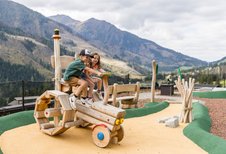 Zwei Kinder spielen auf einem Holztraktor am Abenteuerspielplatz des Familienhotels Almfamilyhotel Scherer mit Bergkulisse im Hintergrund.