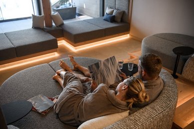 Paar im Zauchenseehof liegt entspannt in grauen Wellness-Liegekissen, in Bademänteln, gemeinsam in ein Magazin vertieft; im Hintergrund moderne Lounge-Sitzflächen mit indirekter Beleuchtung und Blick nach draußen.