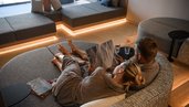 Paar im Zauchenseehof liegt entspannt in grauen Wellness-Liegekissen, in Bademänteln, gemeinsam in ein Magazin vertieft; im Hintergrund moderne Lounge-Sitzflächen mit indirekter Beleuchtung und Blick nach draußen.