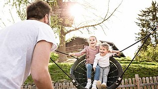 Vater mit zwei Kindern im Last Minute Familienurlaub im Familienhotel. Vater schaukelt die Mädchen auf dem Spielplatz in der Nestschaukel.