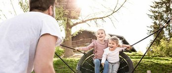 Vater mit zwei Kindern im Last Minute Familienurlaub im Familienhotel. Vater schaukelt die Mädchen auf dem Spielplatz in der Nestschaukel.