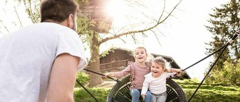 Vater mit zwei Kindern im Last Minute Familienurlaub im Familienhotel. Vater schaukelt die Mädchen auf dem Spielplatz in der Nestschaukel.