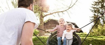 Vater mit zwei Kindern im Last Minute Familienurlaub im Familienhotel. Vater schaukelt die Mädchen auf dem Spielplatz in der Nestschaukel.
