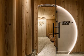 Stimmungsvoll beleuchtete Infrarotkabine im Zauchenseehof mit organisch geformten Liegen, Holzverkleidung und rundem LED-Lichtbogen an der Eingangstür.