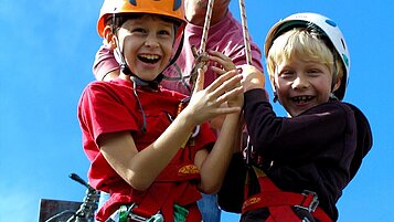 Zwei Jungs hängen mit Kletterausrüstung an einer Seilrutsche und freuen sich auf das Abenteuer.