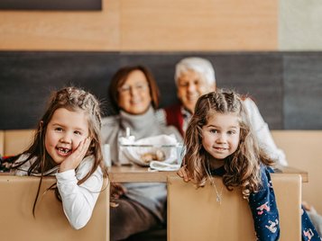Zwei Mädchen lehnen lächelnd über eine Sitzbank, im Hintergrund sitzen die Großeltern am Restauranttisch im AIGO Welcome Family.