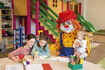 Kinder malen in der liebevollen Kinderbetreuung des Familienhotels Alpenhotel Kindl in Tirol. Zu Besucht ist Clown Happy, das Maskottchen des Familienhotels.