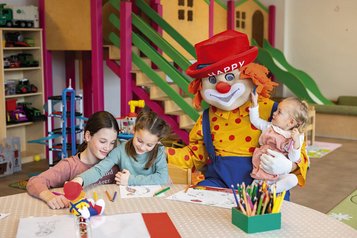 Kinder malen in der liebevollen Kinderbetreuung des Familienhotels Alpenhotel Kindl in Tirol. Zu Besucht ist Clown Happy, das Maskottchen des Familienhotels.